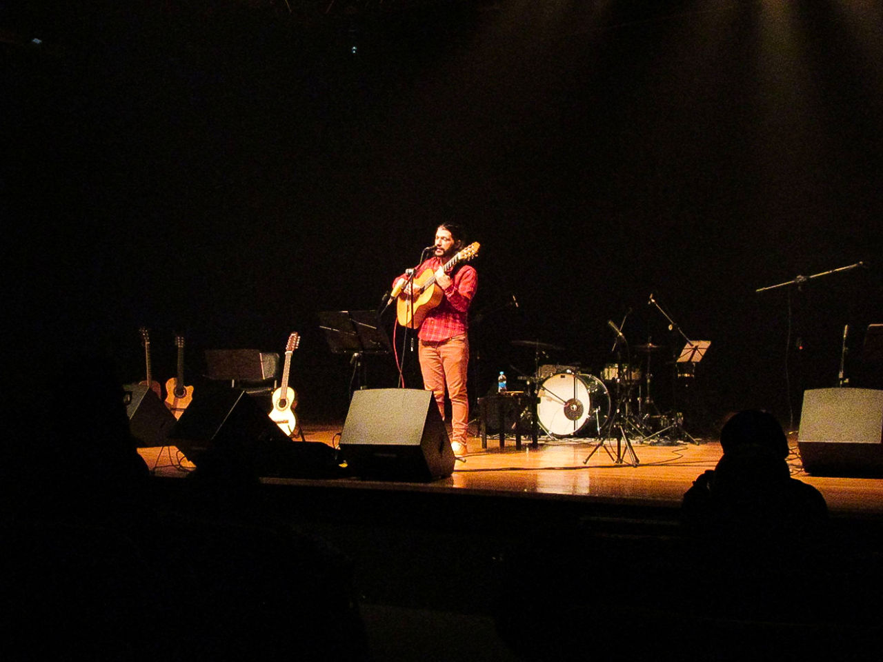 Cantor durante show, com violão na mão, outros instrumentos no palco e luz focada