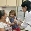 Enfermeira aplica vacina em menina que está no colo da mãe