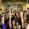 Foto noturna na Praça do Coreto, com a Matriz ao fundo e bailarinos em intervenção de flashmob com as mãos para o alto