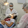 Paciente deitada em cadeira enquanto dentista faz procedimento com assistente ao lado