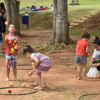 Três crianças brincando de bolas e bambolês, sob árvores e com a supervisão de monitora