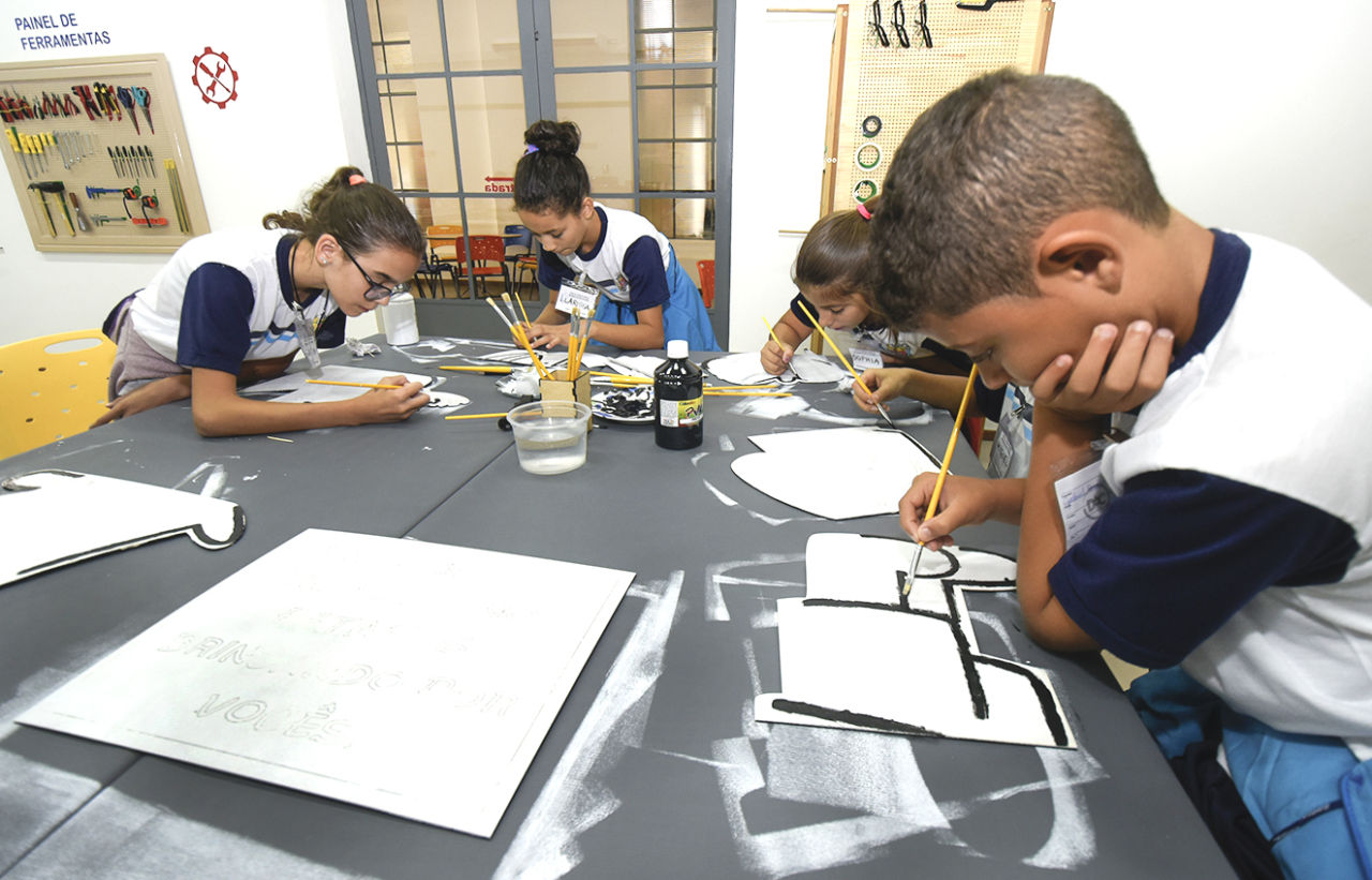 Crianças pintam com pincel placas de madeira, sobre mesa no FAB LAB