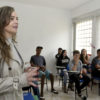Sala de aula com estudantes e mulher dando explicação, voltada para a direção da lousa da sala