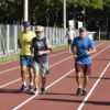 Quatro pessoas correm em pista de atletismo em dia de sol