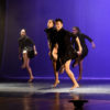 Bailarinos com figurino todo preto, em apresentação no palco do Polytheama, com luzes roxas ao fundo