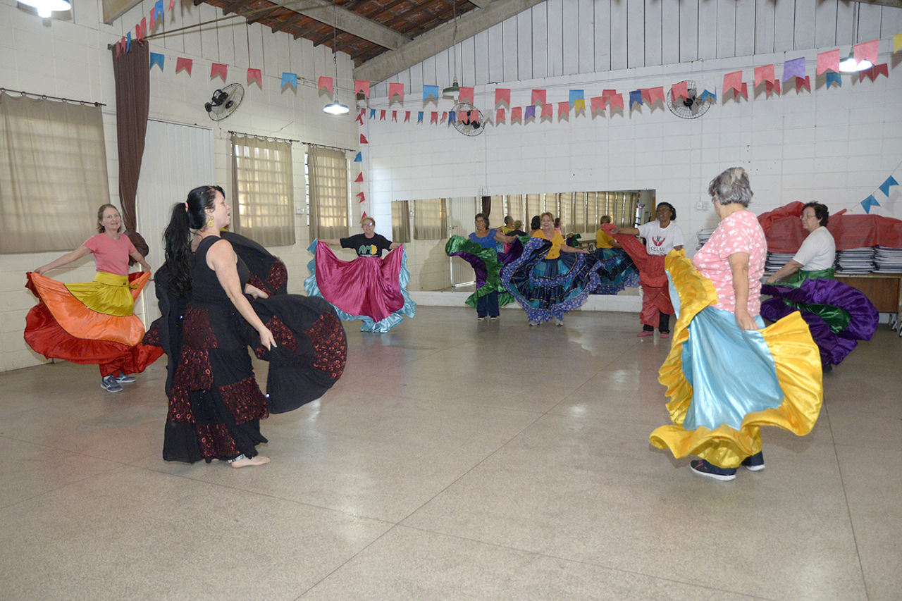 Senhoras fazendo coreografias com saias coloridas, em salão com espelho e decoração com bandeiras juninas coloridas