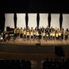 Palco iluminado do teatro Polytheama, com cantores crianças e adultos e pianista, com primeiras filas da plateia à vista