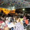 Foto noturna, com pessoas comendo e conversando, sentadas sob uma tenda, com decoração de bandeiras juninas coloridas