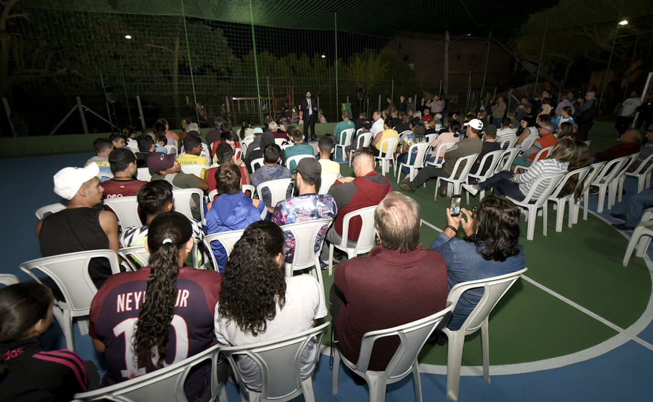 Pessoas sentadas em cadeiras em quadra poliesportiva, à noite, com o prefeito à frente