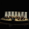 Foto do teatro Polytheama, somente com o palco e musicista da Orquestra iluminados, e com silhueta da plateia no escuro
