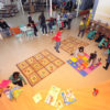 Foto do ambiente de brinquedoteca da Biblioteca, com crianças brincando, mexendo em livros e sobre tapetes interativos