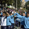 Crianças uniformaizadas e com faixas das cores da bandeira nacional em desfile no meio de uma avenida, com público atrás de gradis