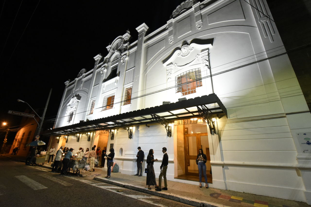 Foto noturna do teatro, com fachada iluminada e pessoas na calçada