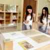 Duas mulheres olham com atenção para instalação de uma exposição, protegida por edoma de vidro, com prateleira com acervos de exposição ao fundo