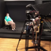Câmera filmadora sobre tripé em primeiro plano, com dois homens sentados, conversando, em posição de entrevistador e entrevistado