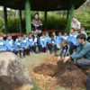 Grupo de crianças sentadas, enquanto uma outra criança ajuda um adulto a plantar uma árvore