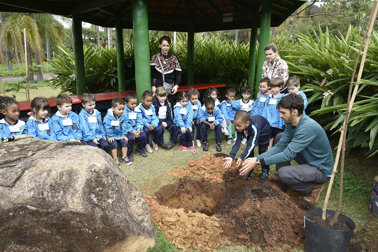 Grupo de crianças sentadas, enquanto uma outra criança ajuda um adulto a plantar uma árvore