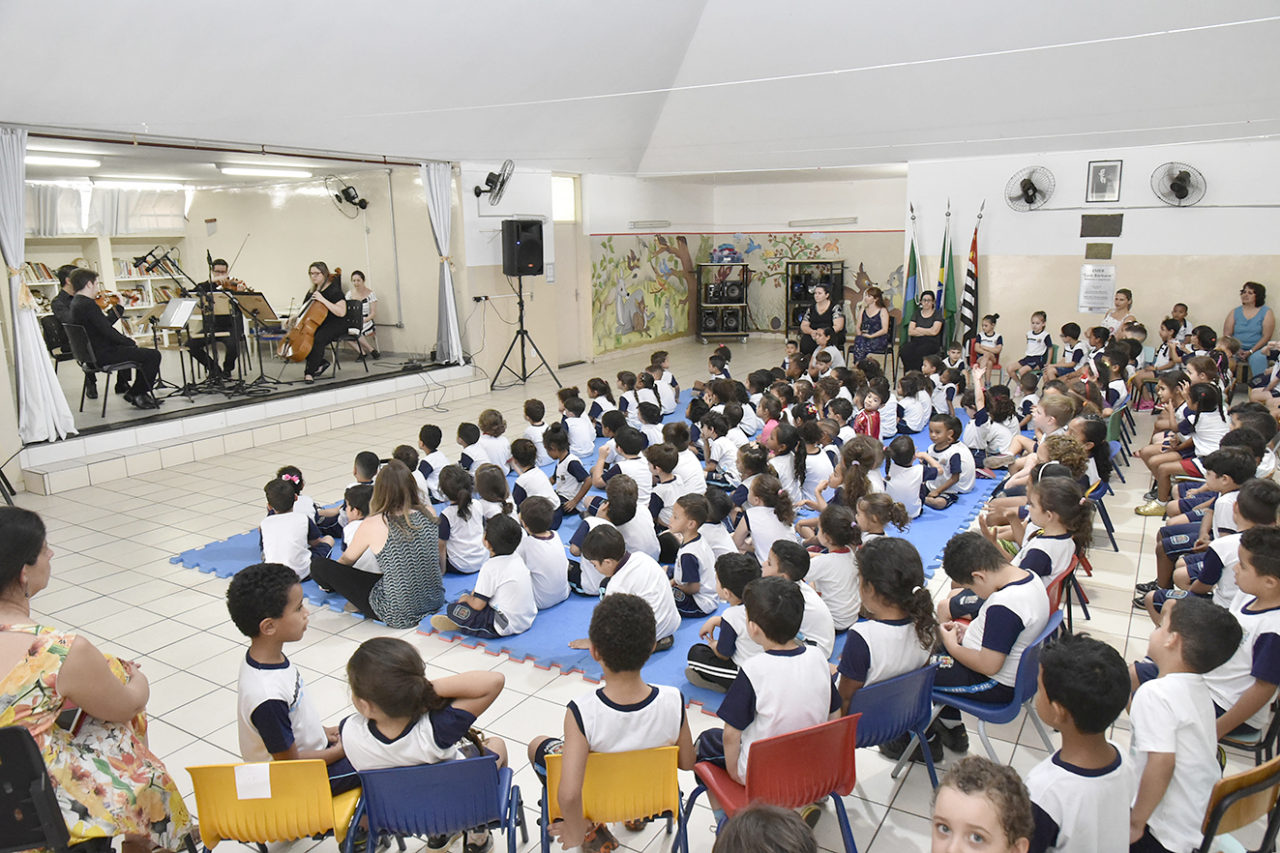 Crianças sentadas no chão e em cadeiras, assistindo a um grupo de músicos