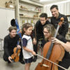 Criança uniformizada segura o arco de um violoncelo, com a atenção de músicos