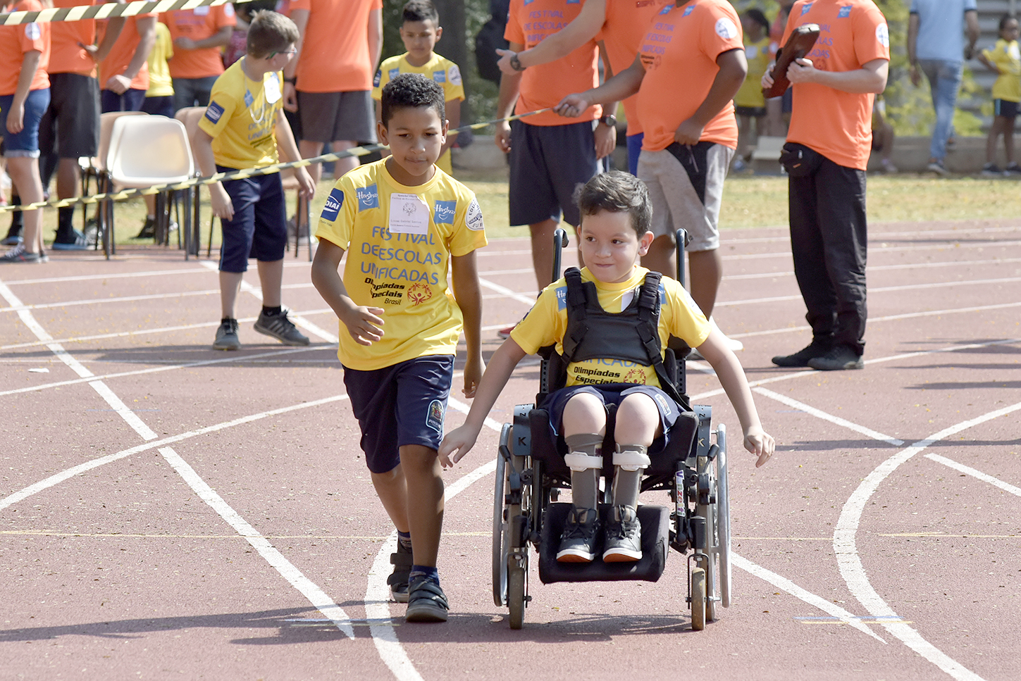Em primeiro plano, criança correndo ao lado de uma criança portadora de deficiência física. Ao fundo voluntários observando o desenvolvimento da atividade