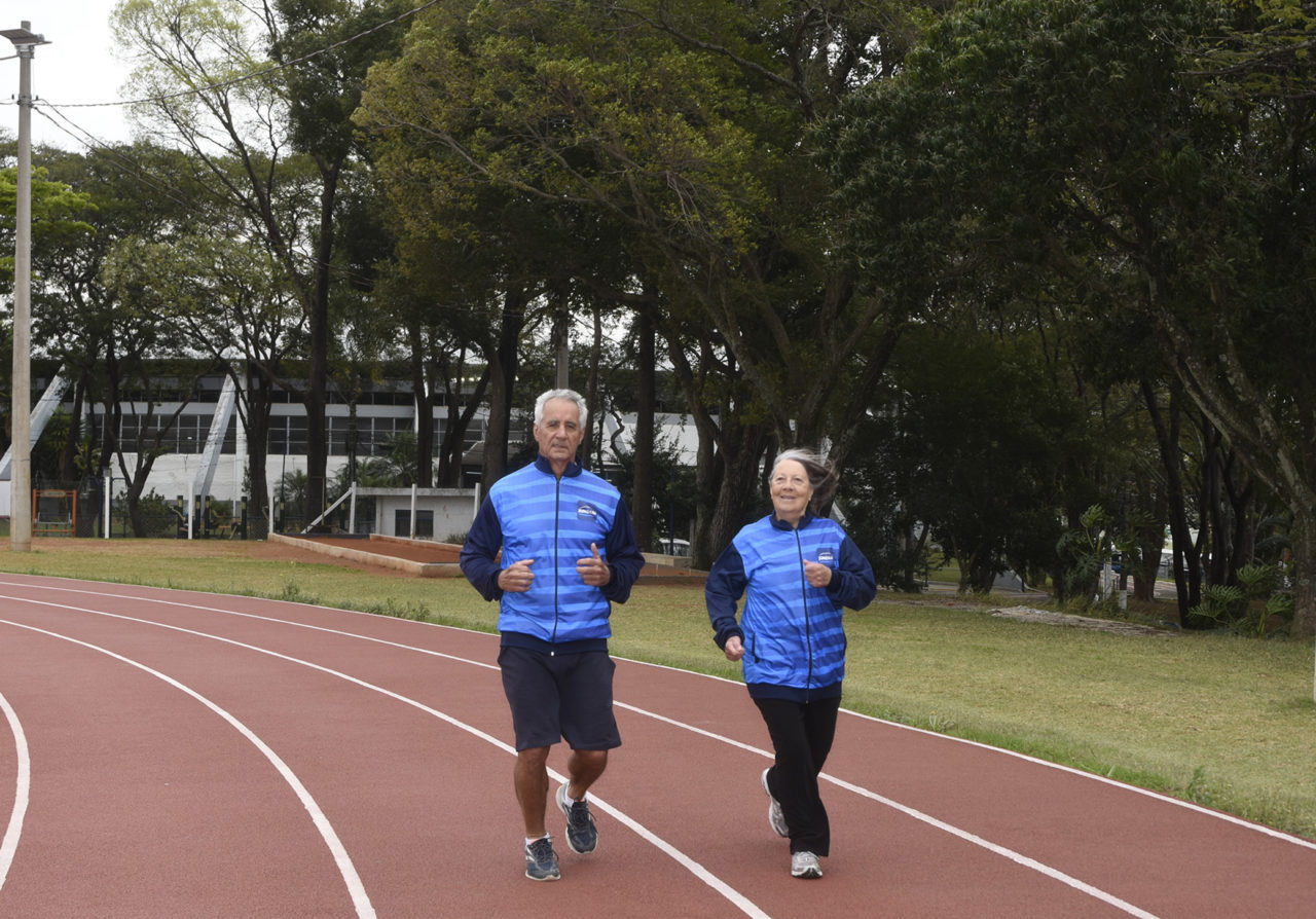 Um homem e uma mulher uniformizados correndo em pista de atletismo, com ginásio ao fundo