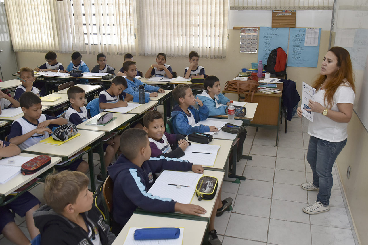 Crianças uniformizadas sentadas em sala de aula, enquanto professora mostra um material
