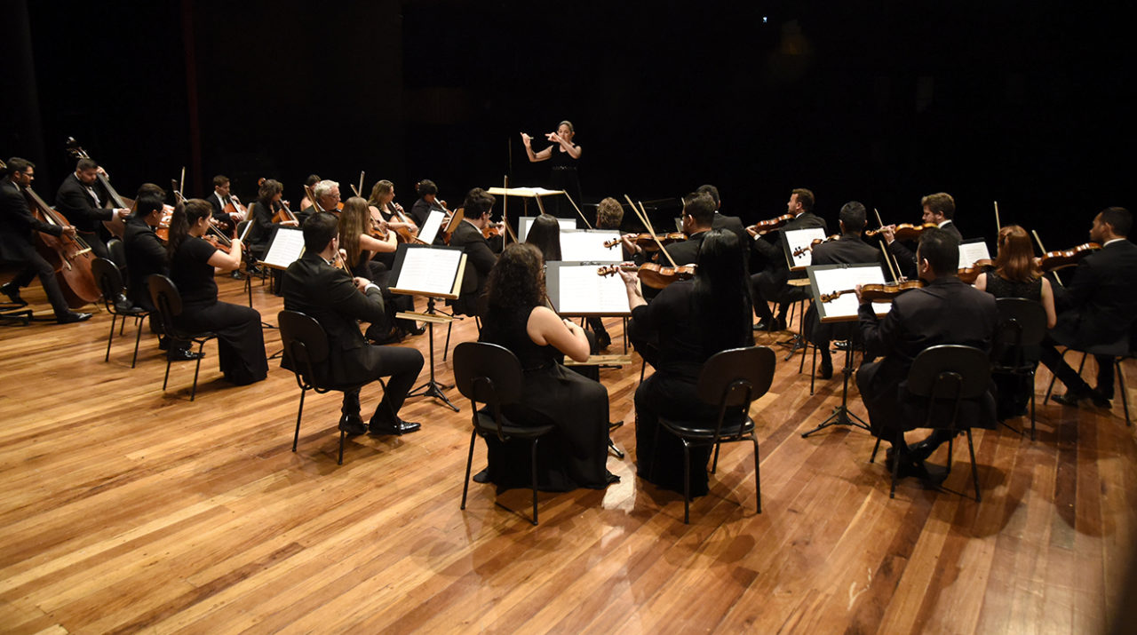 Concerto da Orquestra, com músicos sentados e maestrina ao fundo regendo