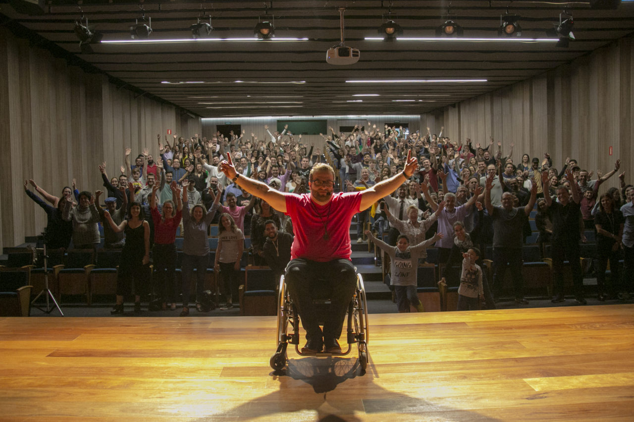 Foto posada de homem sentado em cadeira de rodas, com os braços abertos, e plateia ao fundo 