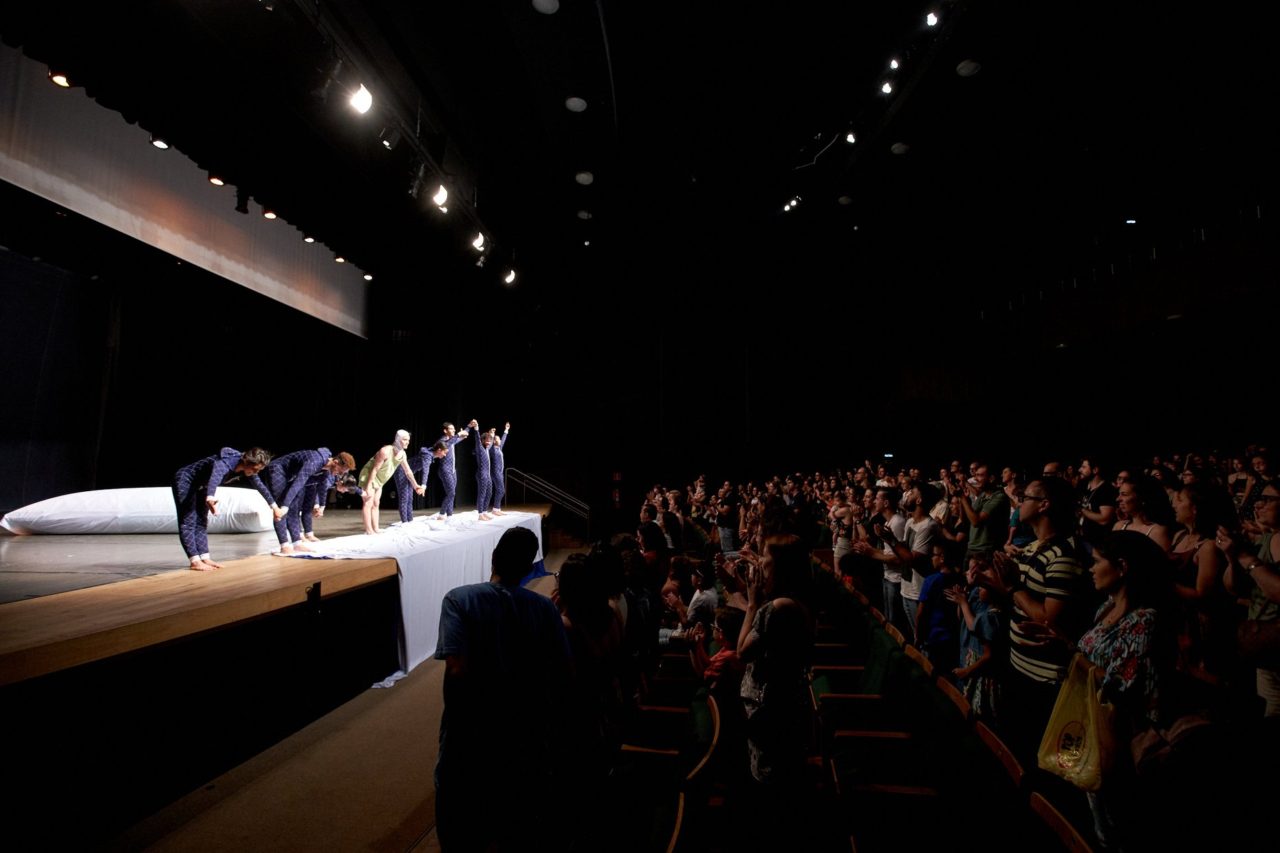 Foto lateral de teatro, com bailarinos no palco para cumprimentos e plateia aplaudindo