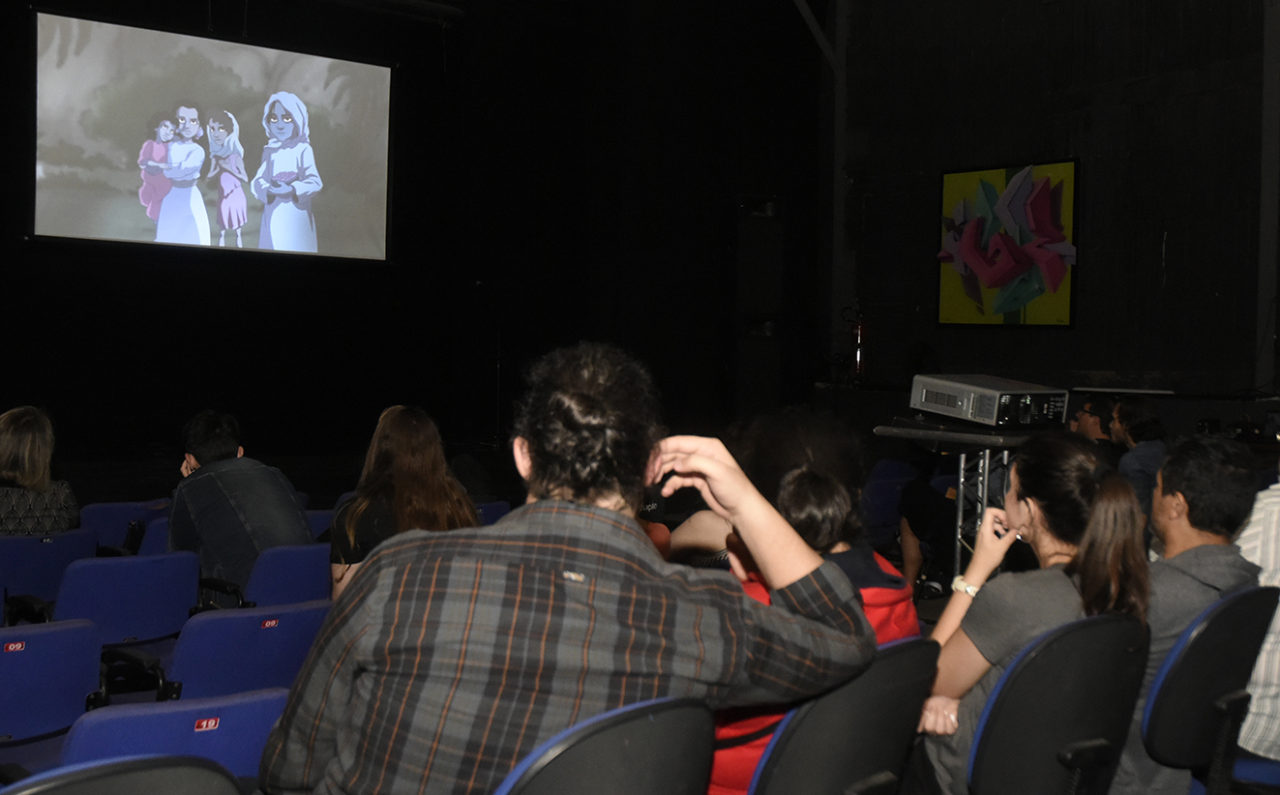 Pessoas sentadas em sala escura, asssistindo a filme sendo projetado em telão branco