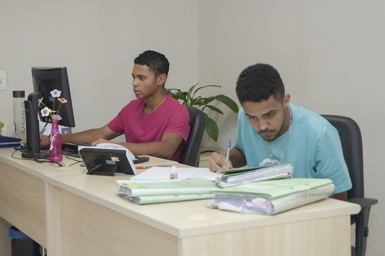 Dois jovens sentados , um trabalhando no computador e outro escrevendo em caderno e manuseando processos