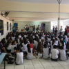 pátio de escola, com crianças sentadas no chão assistindo a músicos