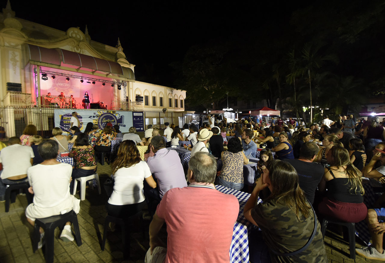Foto noturna da praça com coreto ao fundo e plateia sentada assistindo à apresentação