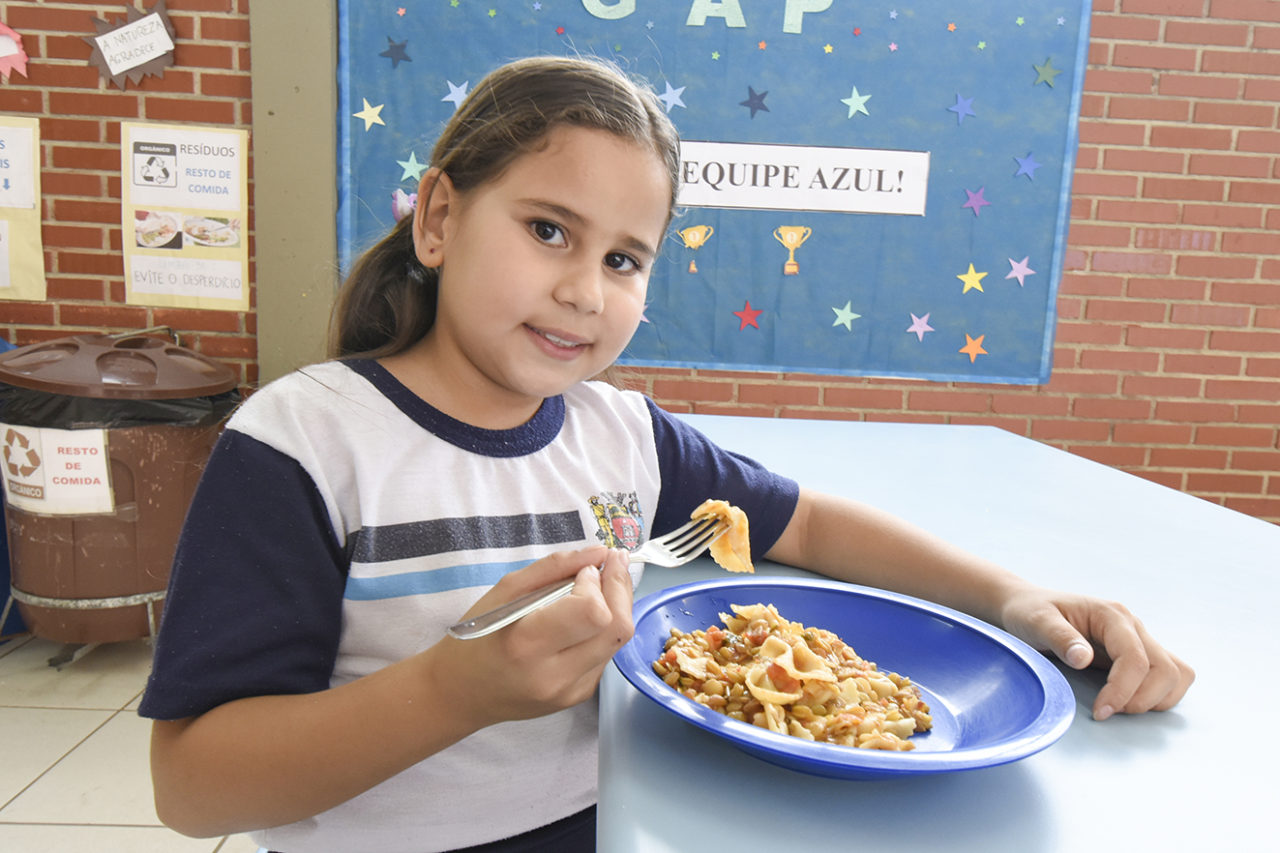 Garotinha estudante comendo macarrão com garfo na mão e prato na mesa