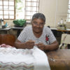 Mulher olhando para a foto enquanto faz crochê e bordado em tecido apoiado em mesa, com janelas e máquinas de costura ao fundo