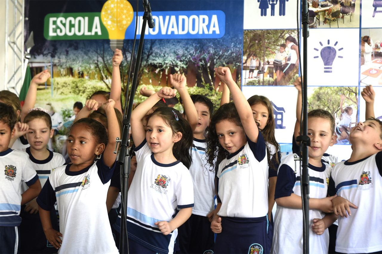 Crianças com uniforme escolar cantando em frente a painel 