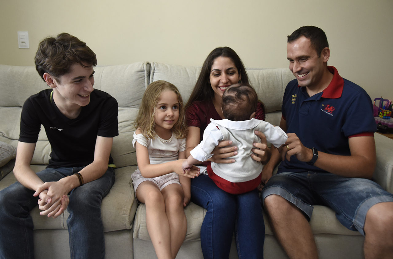 Crianças e adultos sentados em sofá, olhando para bebê de colo, com um bebê de colo, de costas 