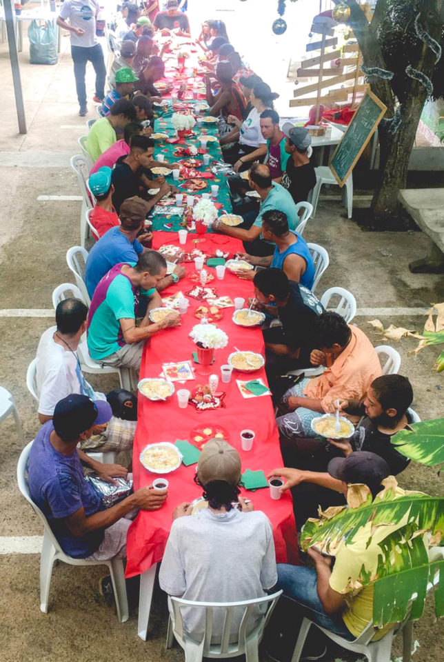 Foto de pessoas sentadas e almoçando ao redor de mesa comprida