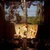 Palco de teatro, com iluminação sobre músicos e atores encenando, com painel luminoso ao fundo