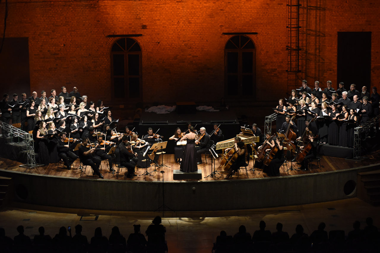 Palco de teatro, com músicos de orquestra e cantores de coral em arquibancadas montadas dos dois lados