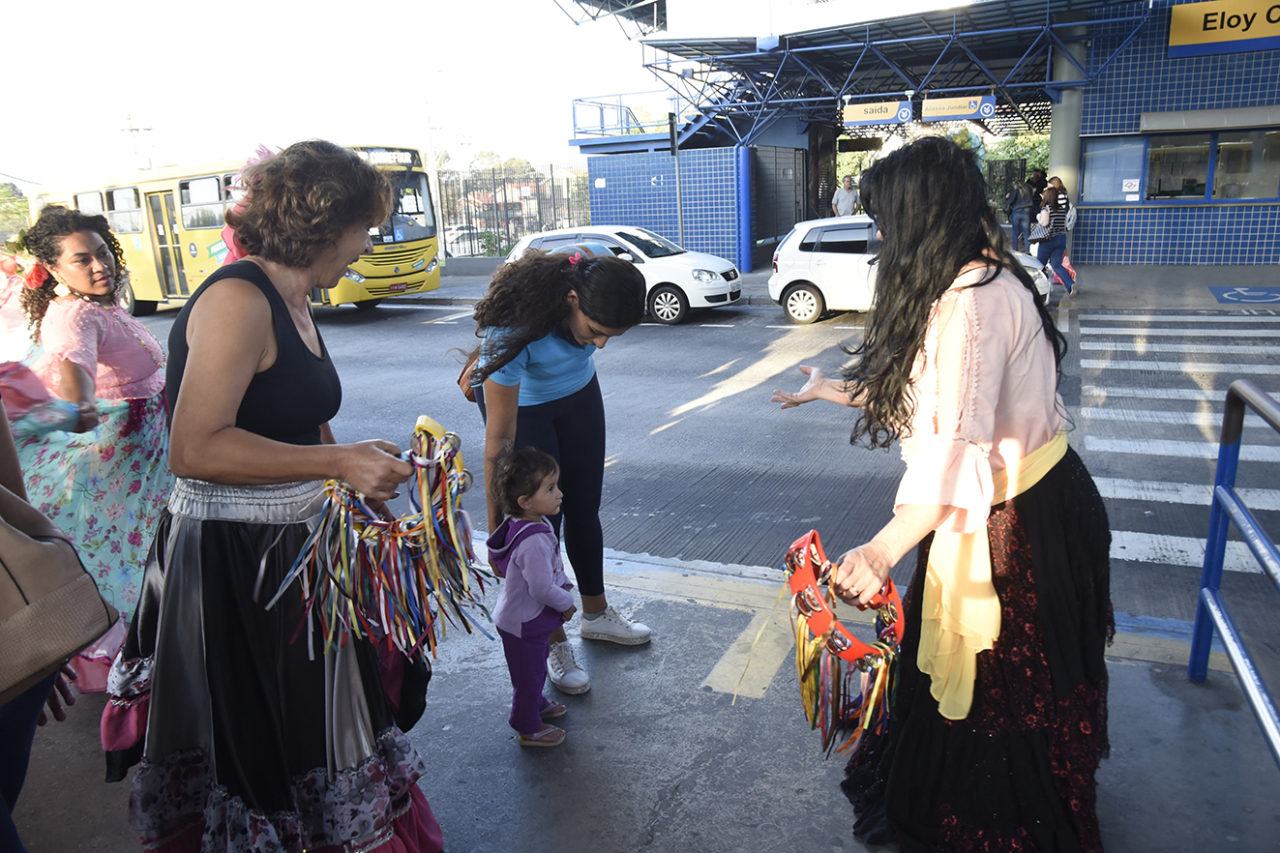 Dançarinas com saias longas e pandeiros coloridos interagindo com criança e mulher em plataforma de terminal de ônibus