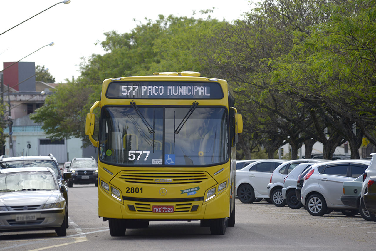 ônibus transitando por via com carros estacionados em ambos os lados, com árvores na calçada