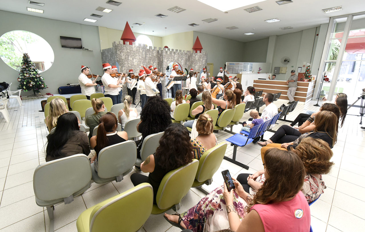 Sala de espera de hospital, com pessoas sentadas, assistindo a uma apresentação de músicos, usando gorros iguais ao do Papai Noel