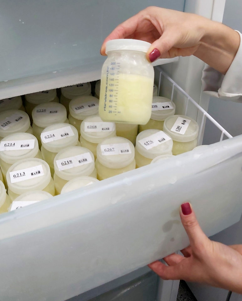 Mão de mulher segura um frasco de leite com gaveta de freezer aberta com mais frascos
