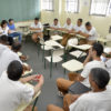 Sala de aula, com pessoas uniformizadas sentadas em círculo, sobre cadeiras escolares
