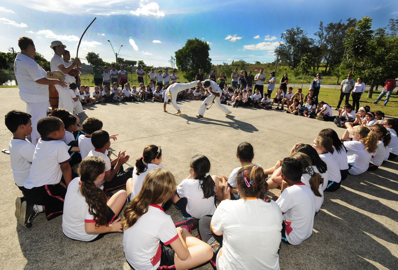 Roda de capoeira, com crianças de uniforme escolar e pessoas com roupa de capoeira