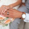 Mãos dos noivos com alianças sobre buquê de flores