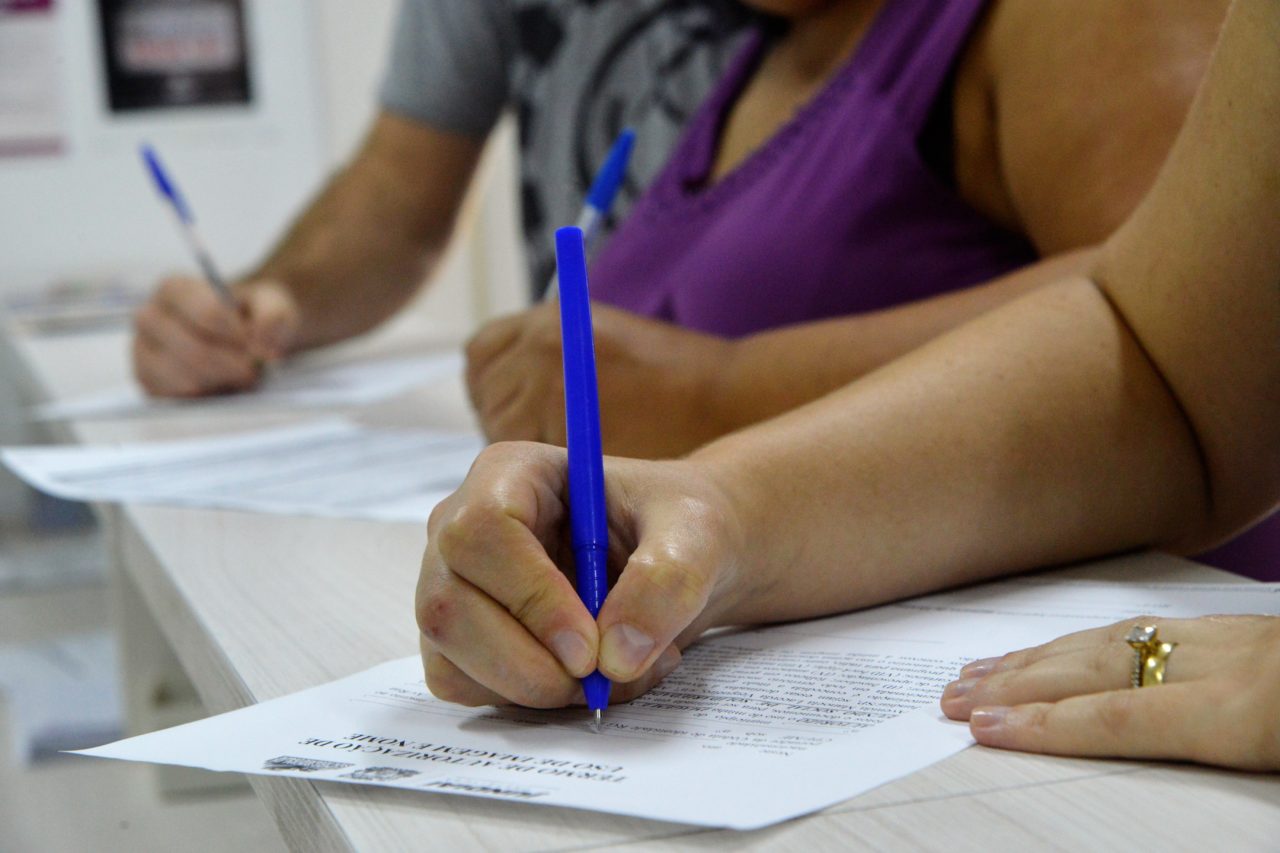Mão de aluna, preenchendo a ficha de inscrição com caneta azul