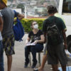 Mulher sentada, usando luvas e máscaras, tomando anotações em prancheta, enquanto conversa com pessoas para serem atendidas
