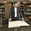 Foto dentro de biblioteca com estantes de livros, homem em pé folheia encadernação em tamanho grande, apoiado sobre mesa